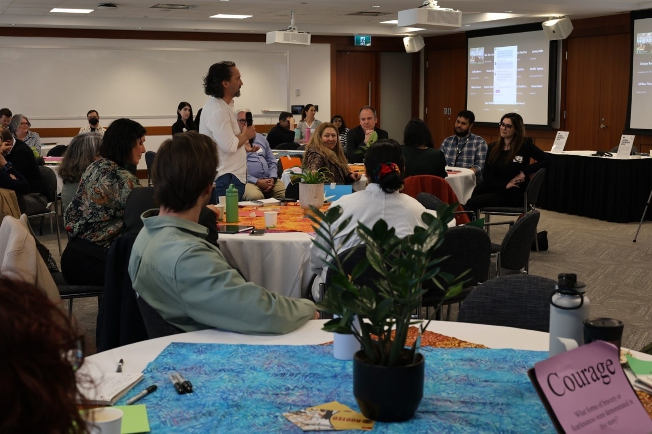A room full of participants all looking towards one participant who is standing and speaking while at a table. Participants are sitting at tables with plants and colourful batik tablecloths on them. There is a pink piece of paper visible on one of the tables that says “Courage” and some smaller text below, as well as a few pieces of colourful paper around the room.