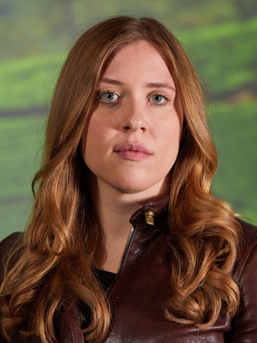 Genevieve is photographed in front of a green landscape. She is wearing a brown leather jacket and has long wavy hair.