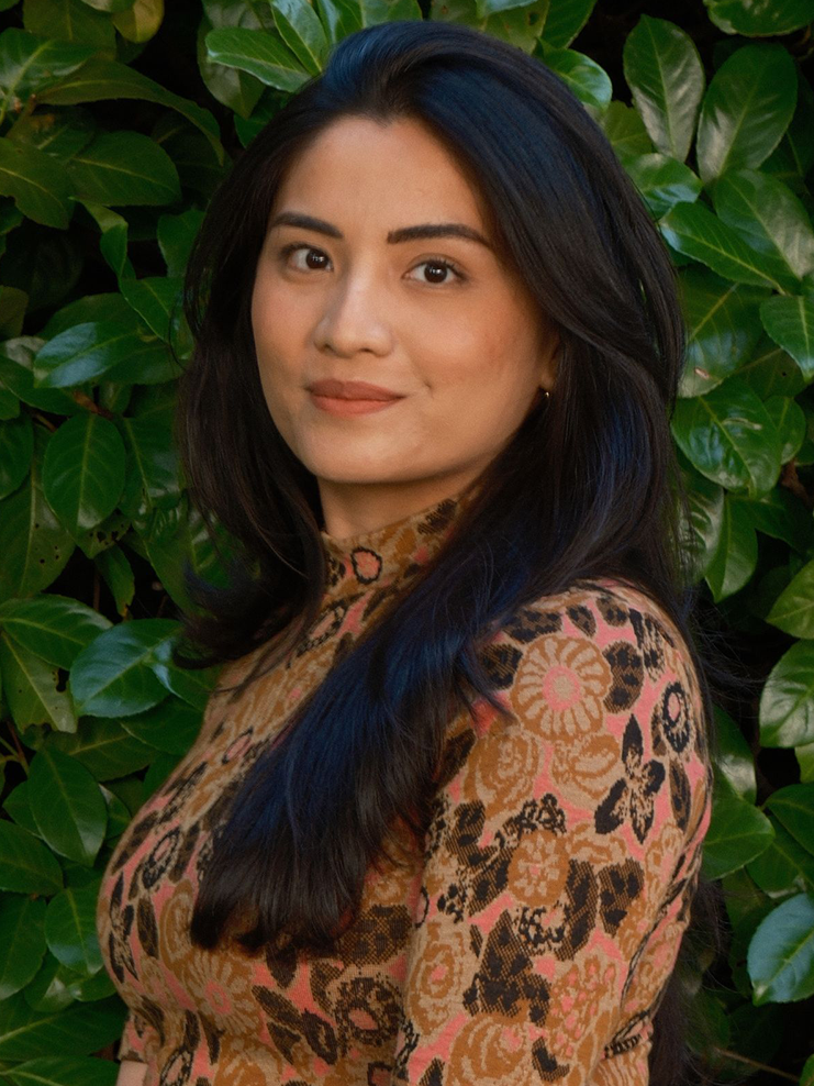 Fiana is photographed outside in front of greenery. She has long dark hair and is wearing a brown patterned shirt.