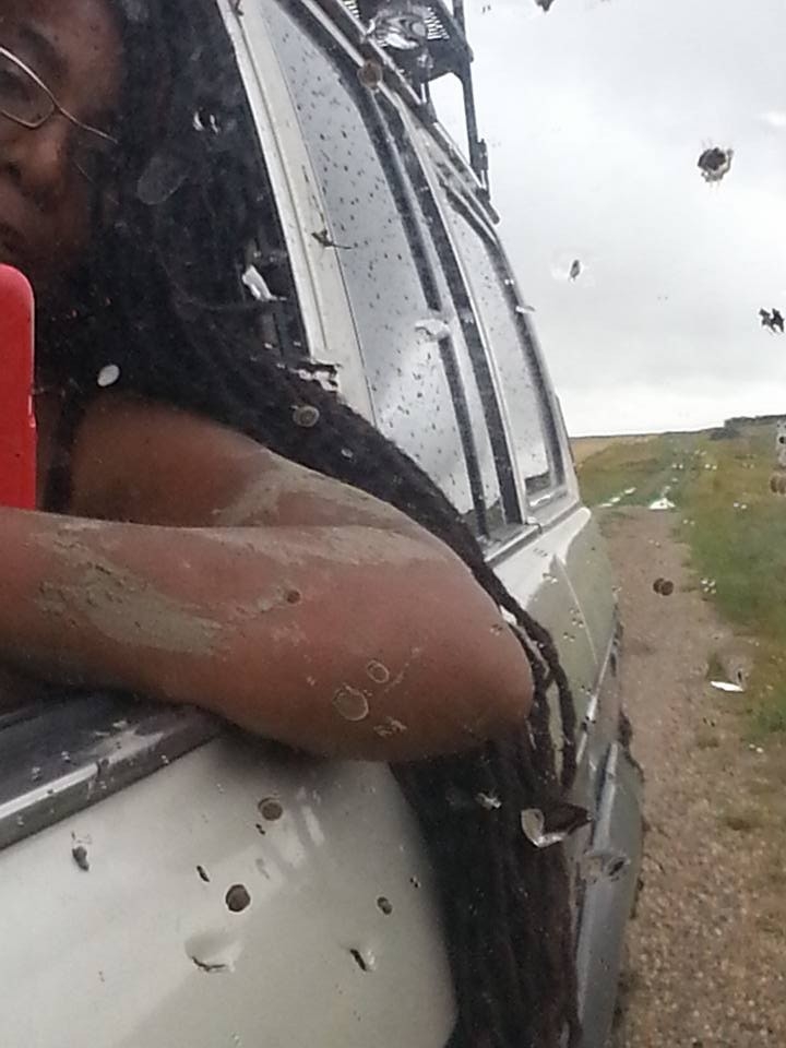 A photo of Nadine in the front seat of a car her arms covered in mud having dug herself out of a field. Nadine has long black dreadlocks and wears glasses.