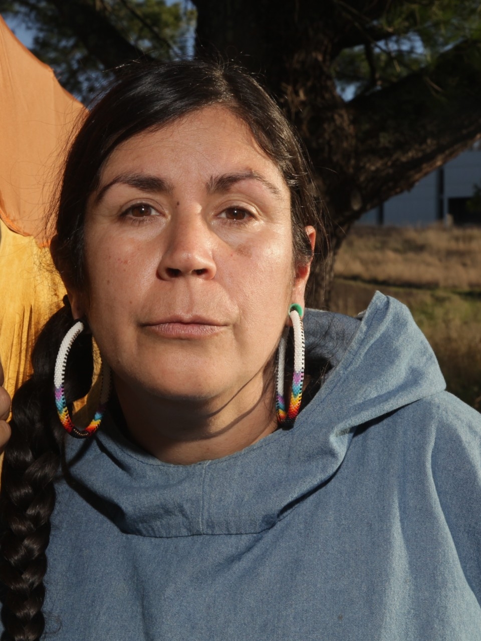 Tania is photographed outside near a woodsy area. She wears large beaded hoop earrings, a grey jacket, and has long brown hair tied into a braid.