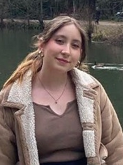A photo of Regina taken in front of a lake. Regina has long brown hair and is wearing a brown coat.