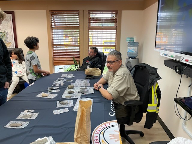 Rudy Reimer sits at a table indoors with artifacts on top for elementary students to observe