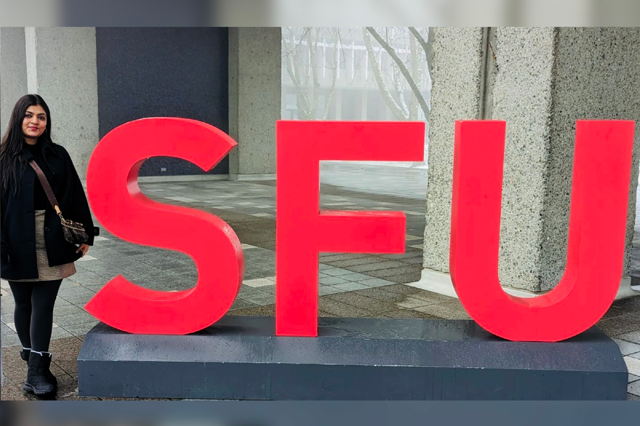 Nidhi Ghelani smiling next to the SFU sign at the AQ 4000 level at SFU Burnaby on a winter day