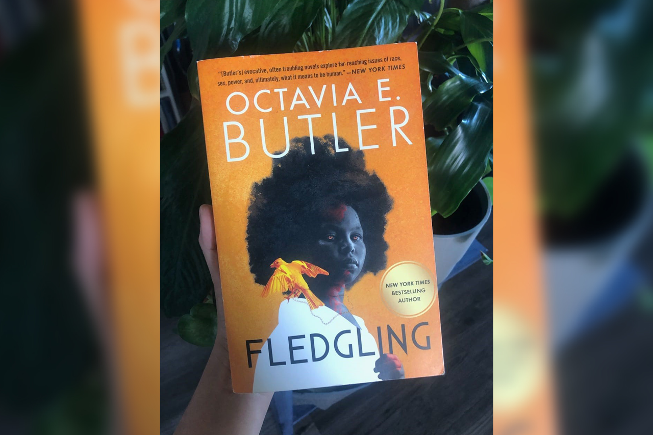 A hand holding a copy of Fledgling by Octavia Butler in front of a plant