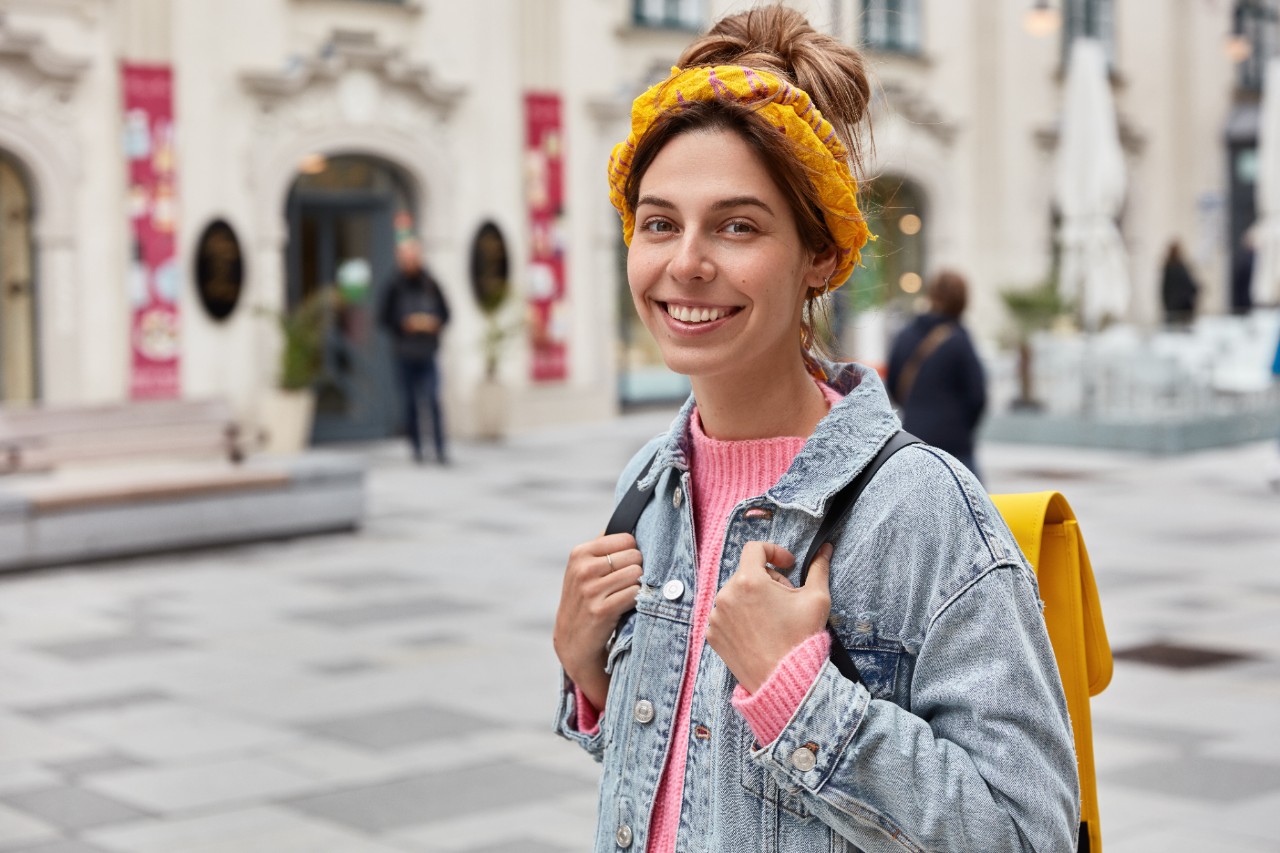Positive optimistic woman has happy expression, wears stylish clothes, has yellow small rucksack on back, wanders across city streets, has carefree expression, covers long distance, explores sights