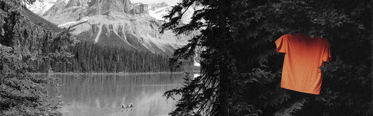 Black and white image of Canada's wilderness, with a lake, mountain, and trees. A bright orange t-shirt is hung from a tree branch, the only colour in the image. 