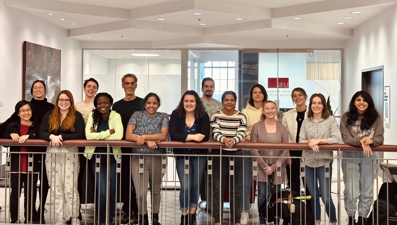 A group of approximately 20 graduate students in the Master of Publishing program standing together. They are in downtown Vancouver, the waterfront and mountains are visible in the background.