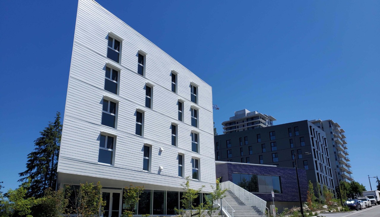 outside picture of Graduate and Family Housing SFU Burnaby Campus. Shows 3 buildings. The West tower is grey, the pavilion in the middle is purple and the East building is a dark grey. Sunny outside