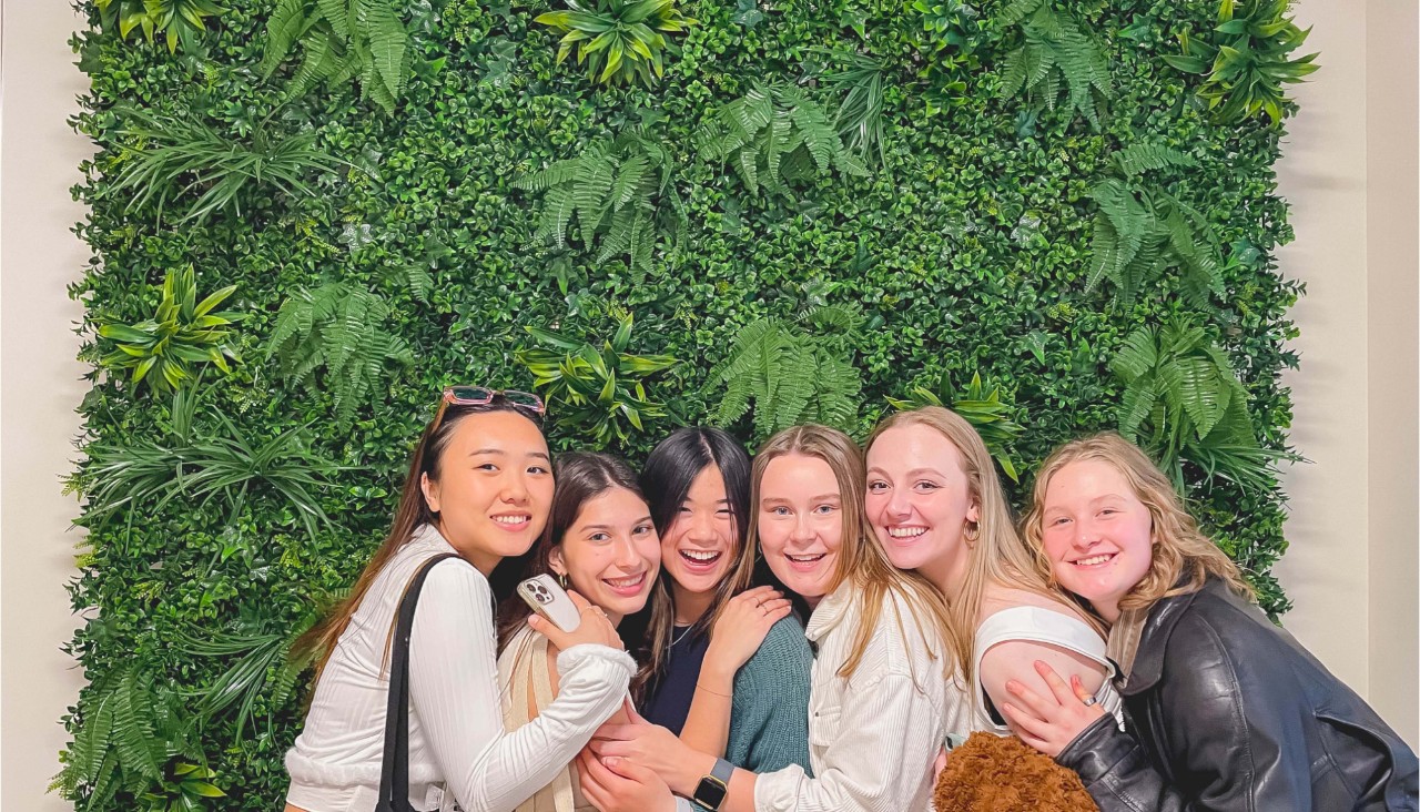 A group of female undergraduate students hanging out at a restaurant standing in front of a green leafy decor