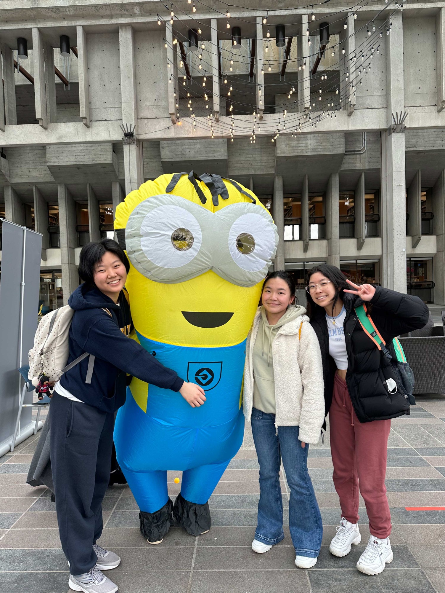 3 students hanging out with a minion costume in front of SFU library smiling. Photo is taken by Alice, a student resident living in Barbara Rae House