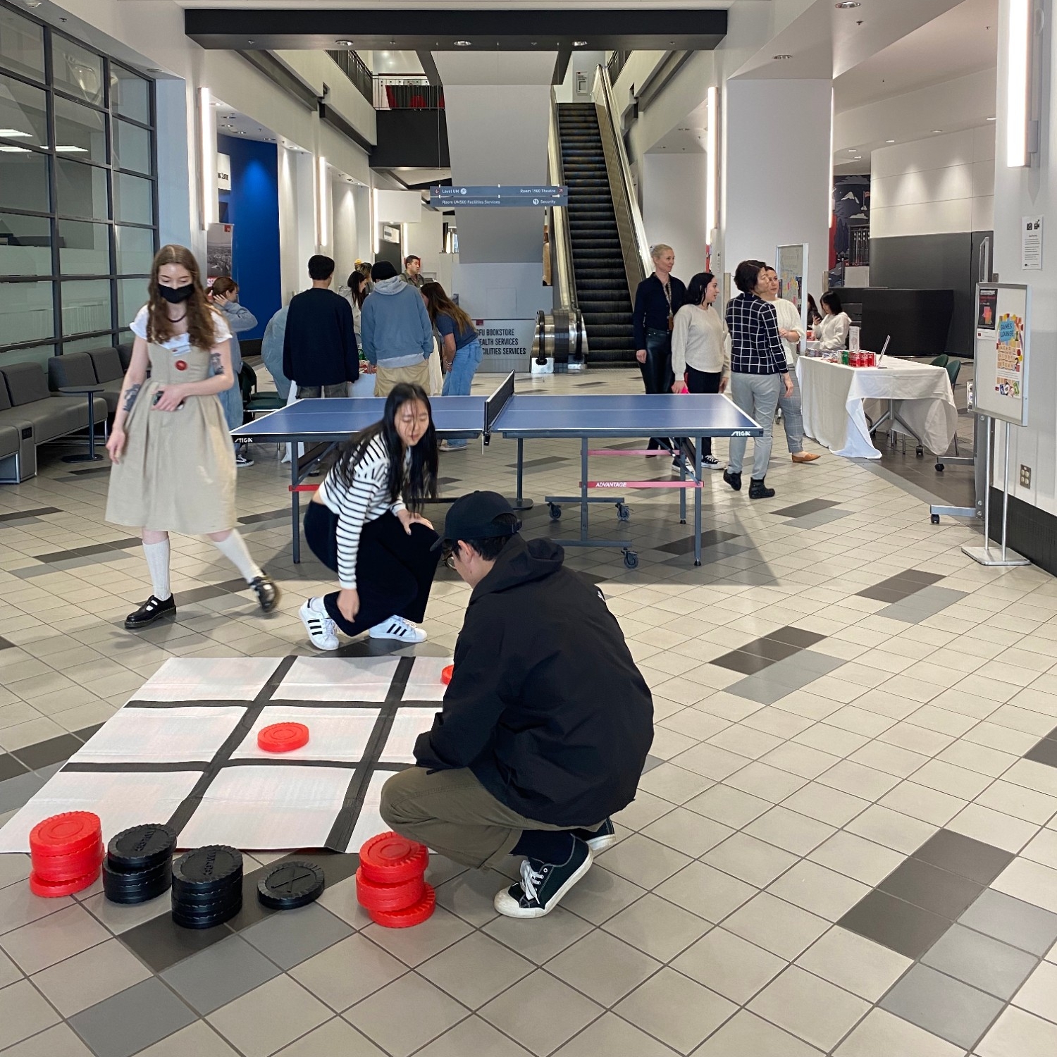 Three students playing giant Checkers with a ping pong table and multiple people in the background at Multilingual Week 2023 at Vancouver campus