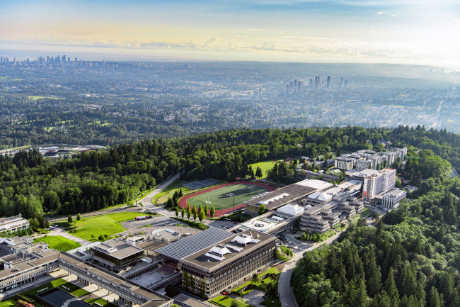An aerial photograph of SFU's Burnaby Campus in the late afternoon with the cities of Burnaby and Vancouver visible in the background