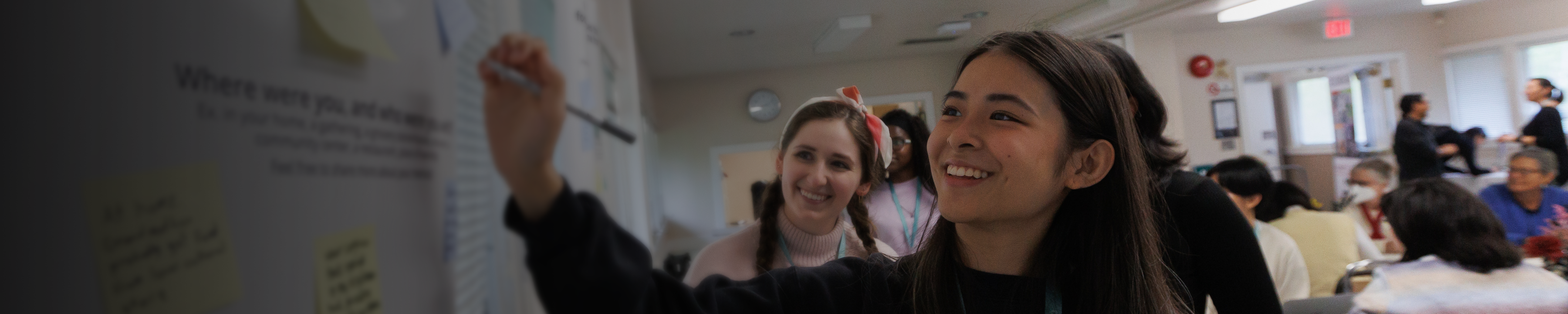 an SFU student with a marker, facing a poster covered in sticky notes with others meeting behind