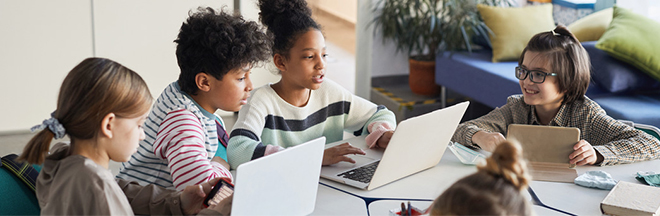 Four kids sitting and communicating at a desk while on their laptops