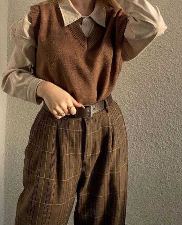 An image of a girl wearing a brown sweater vest on top of a beige shirt, and plaid pants with a belt.