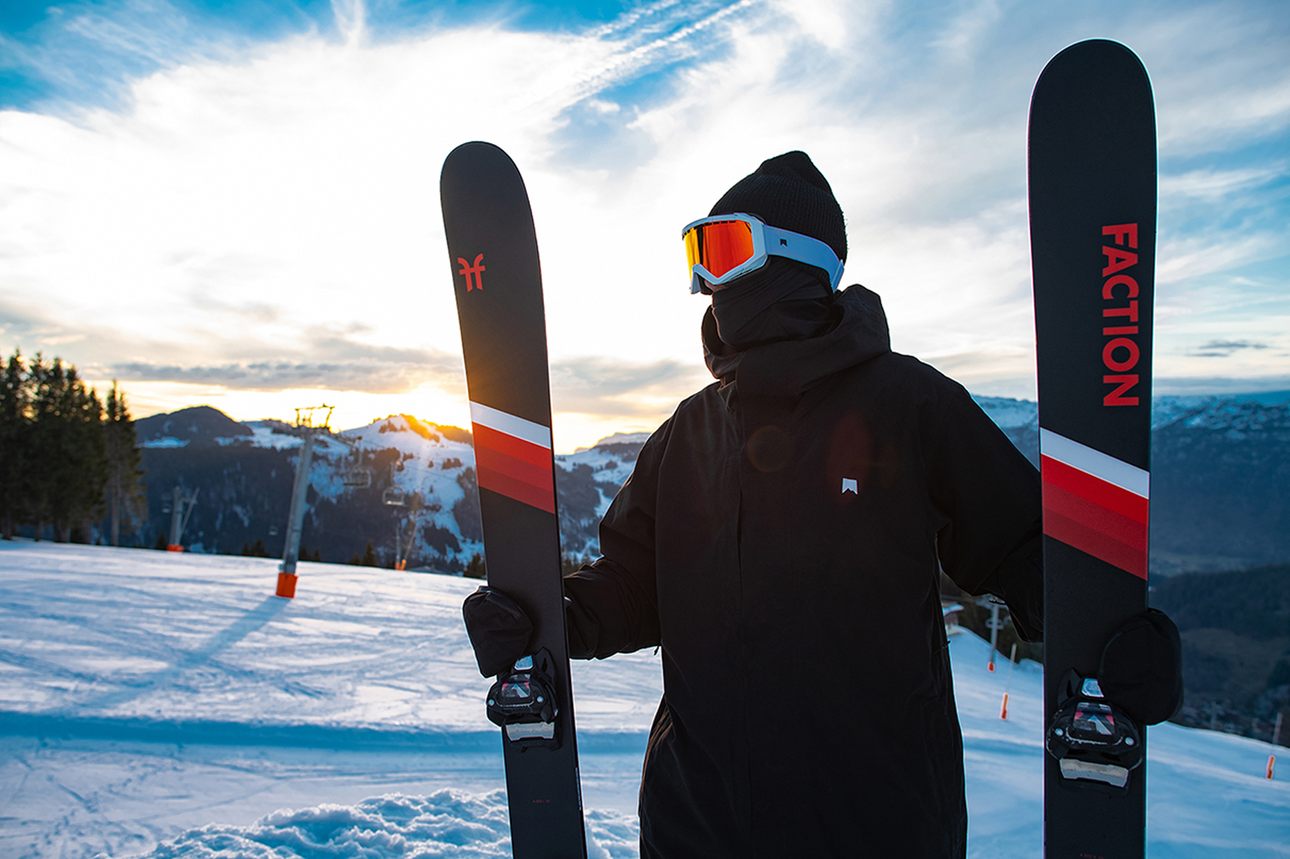 skiier in black and white goggles hold faction skis (that are stuck onto the snow vertically) during sunset 