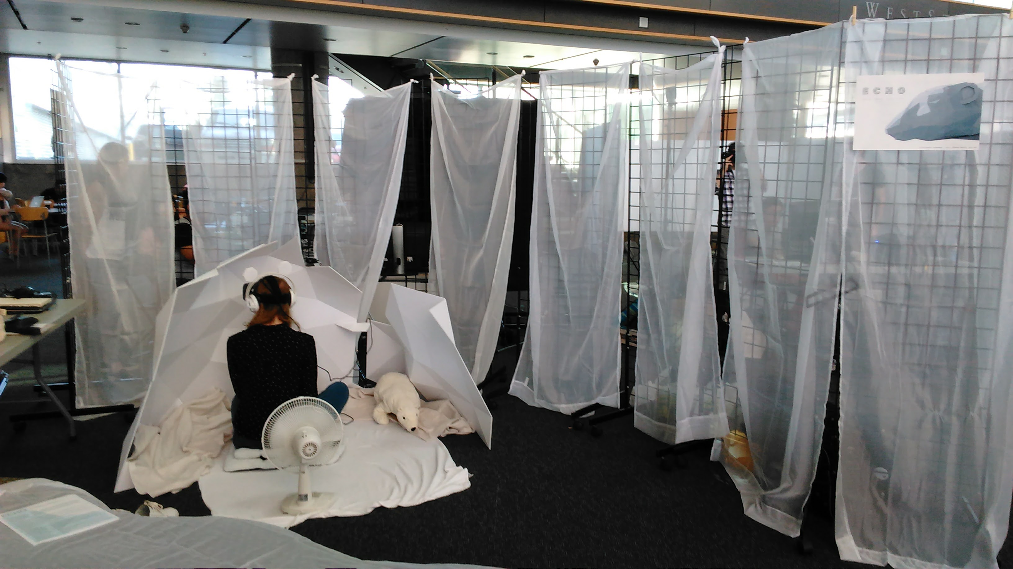 A photograph of Echo's physical installation at the SFU Mezzanine, showing a lady sitting on a white cloth inside a low-poly cave, facing away from the camera, surrounded by grid frames covered in white and transparent fabric