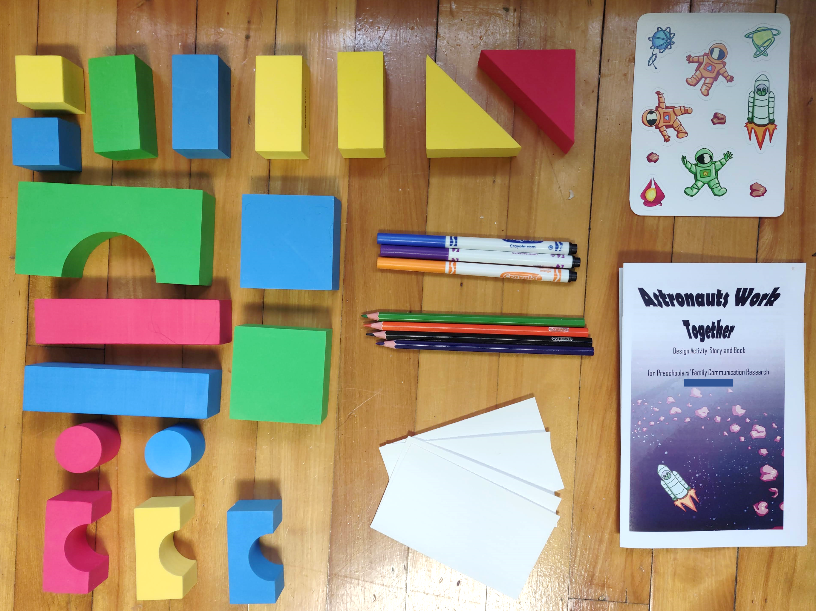 A group of colourful blocks in different shapes (squares, rectangles, triangles, arches, and cylinders) are laid out on a wooden floor. Next to them are 3 markers and 4 coloured pencils in different colours, 4 blank index cards, a sheet of stickers showing astronauts, spaceships, and asteroids, and an activity book titled 'Astronauts Work Together' with an illustration of a green spaceship flying near an asteroid belt.