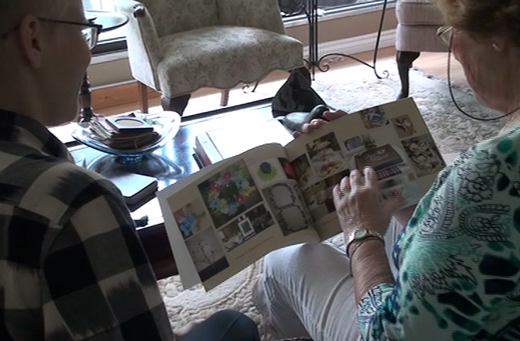 An older person in a living room holding a photobook and pointing to a particular picture while a young person observes.