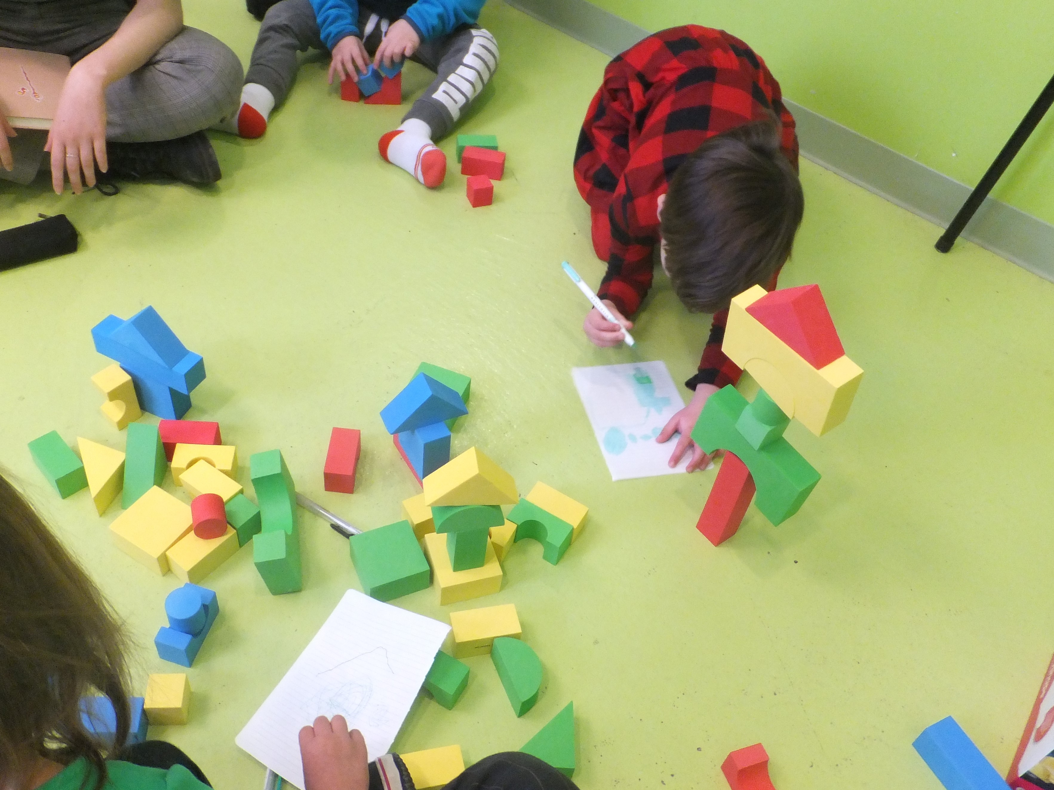 Three children and one adult sitting in a circle around colorful blocks, pages with drawings, and markers.