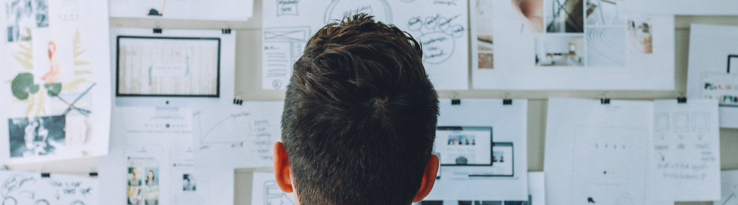 Banner image of the back of a man's head as he looks at a wall covered in papers